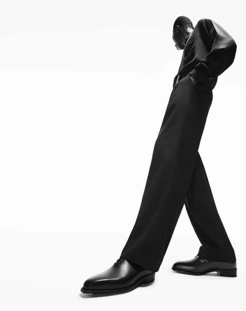 Monochrome fashion portrait of an African-American man in a relaxed pose wearing an all-black suit with leather dress shoes on white.
