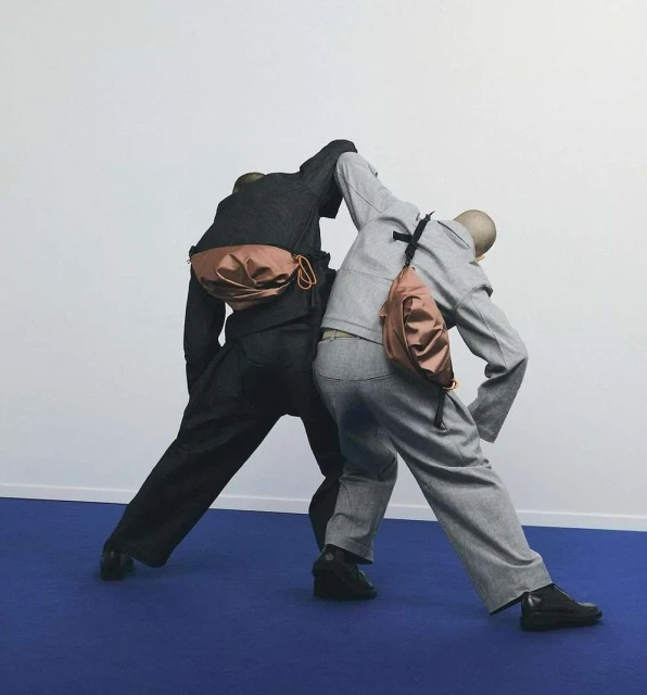Two mannequin torsos wear gray and black suits, accessorized with pink fanny packs in a studio setting against a blue floor.