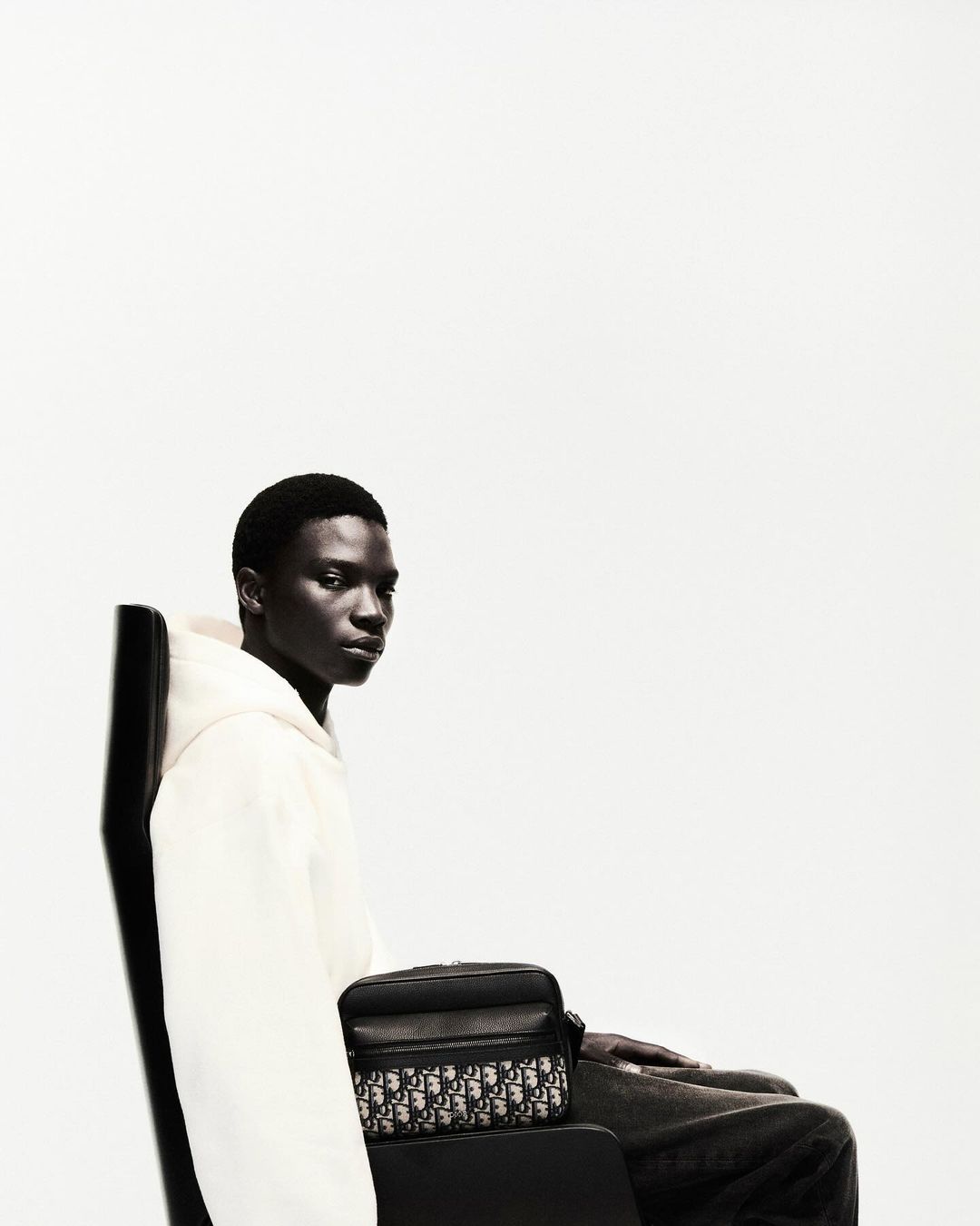 Dark-skinned man wearing white hoodie sitting in a black chair with a black patterned cross-body bag against a white background.