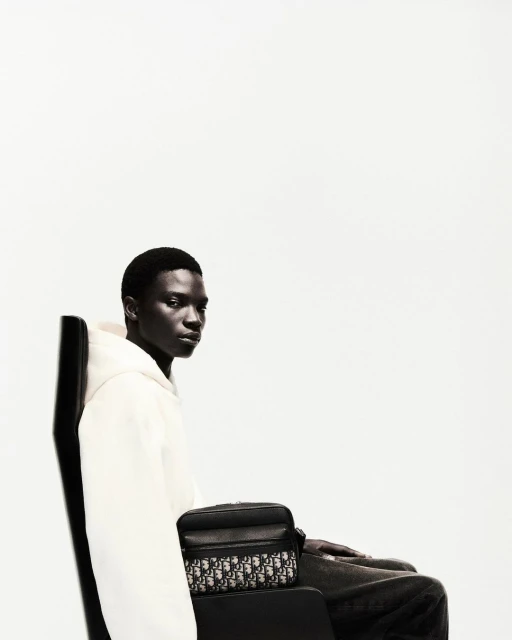 Dark-skinned man wearing white hoodie sitting in a black chair with a black patterned cross-body bag against a white background.