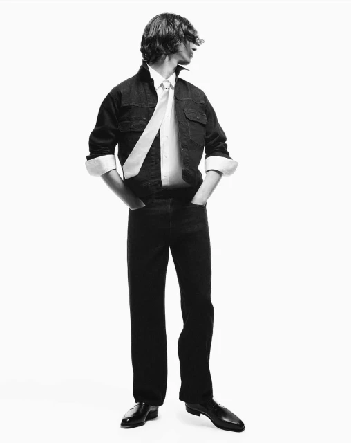 Monochrome portrait of a young man in a denim jacket, white shirt and black pants, looking over his shoulder in a bright, clean photo studio.