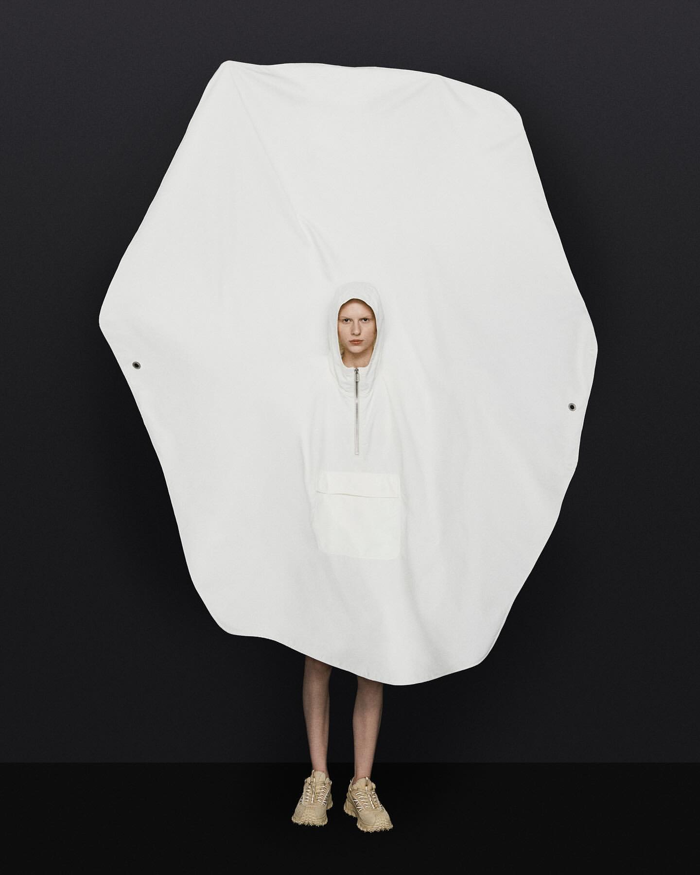 Woman in a large white hooded poncho standing against a black background, emphasizing a minimalist fashion statement and studio lighting.