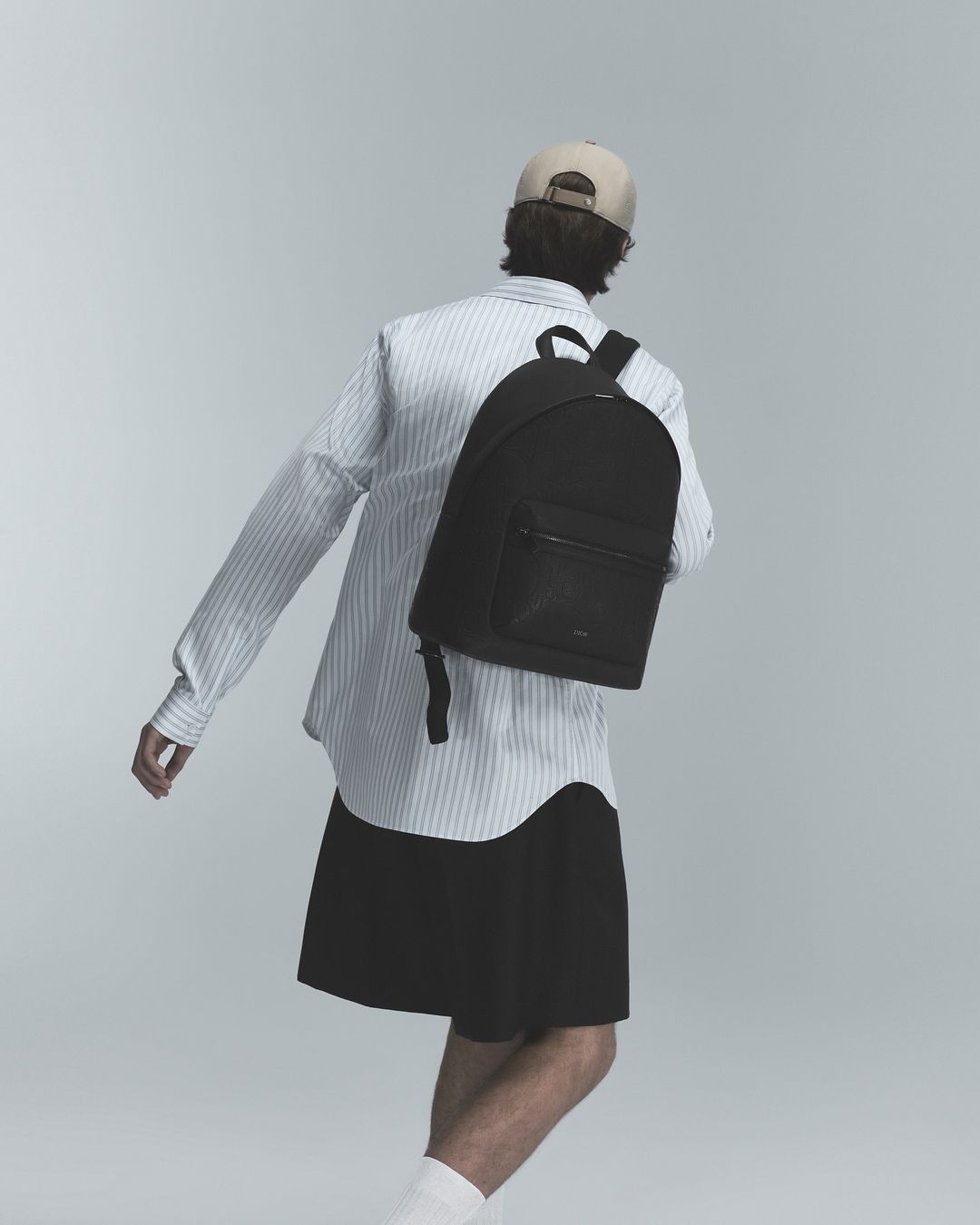 Man wearing a striped shirt, cap, and black backpack seen from behind against a neutral grey studio background