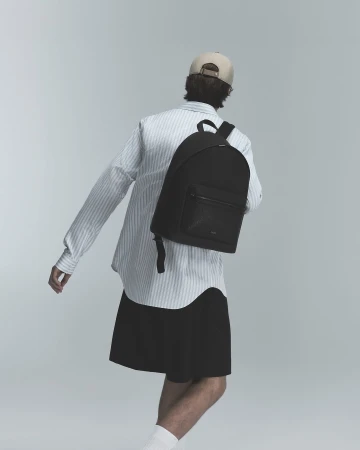 Man wearing a striped shirt, cap, and black backpack seen from behind against a neutral grey studio background