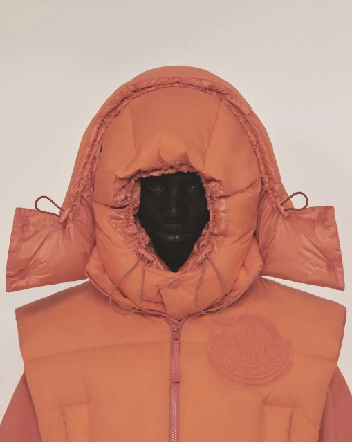 Person wearing a hooded, coral-colored puffer vest with the brand logo on the left side, set against a plain light background.
