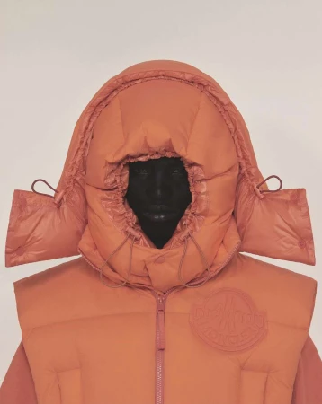 Person wearing a hooded, coral-colored puffer vest with the brand logo on the left side, set against a plain light background.