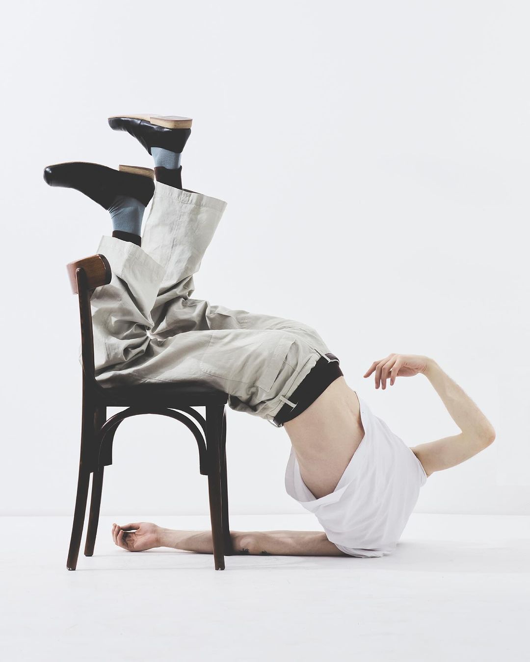Man in a white tank top and beige pants lies upside down with his torso resting on a brown chair in a bright white studio.