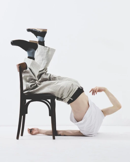 Man in a white tank top and beige pants lies upside down with his torso resting on a brown chair in a bright white studio.