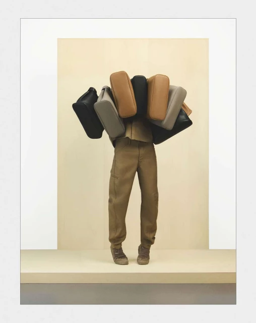 A person is wearing a khaki outfit and carrying a collection of handbags in varying neutral tones against a tan backdrop.