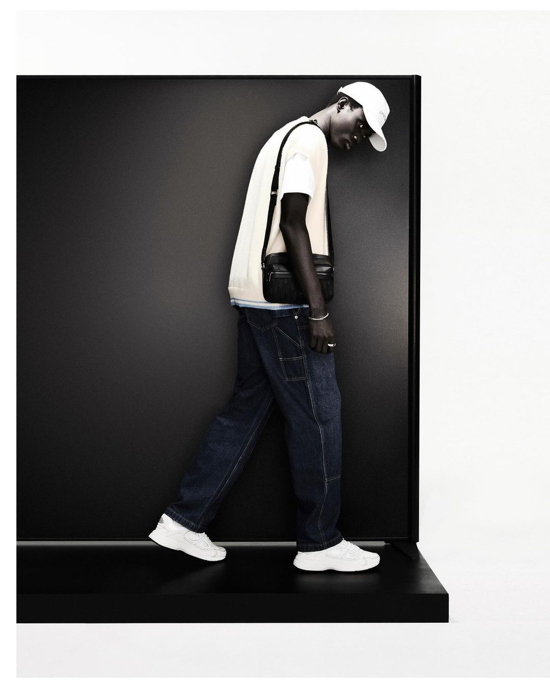 Fashion shot of a dark-skinned man in jeans, t-shirt, baseball cap, and white sneakers, posing in front of a black and white background.