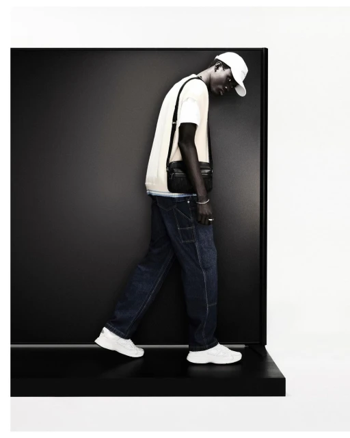 Fashion shot of a dark-skinned man in jeans, t-shirt, baseball cap, and white sneakers, posing in front of a black and white background.