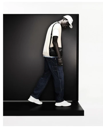 Fashion shot of a dark-skinned man in jeans, t-shirt, baseball cap, and white sneakers, posing in front of a black and white background.