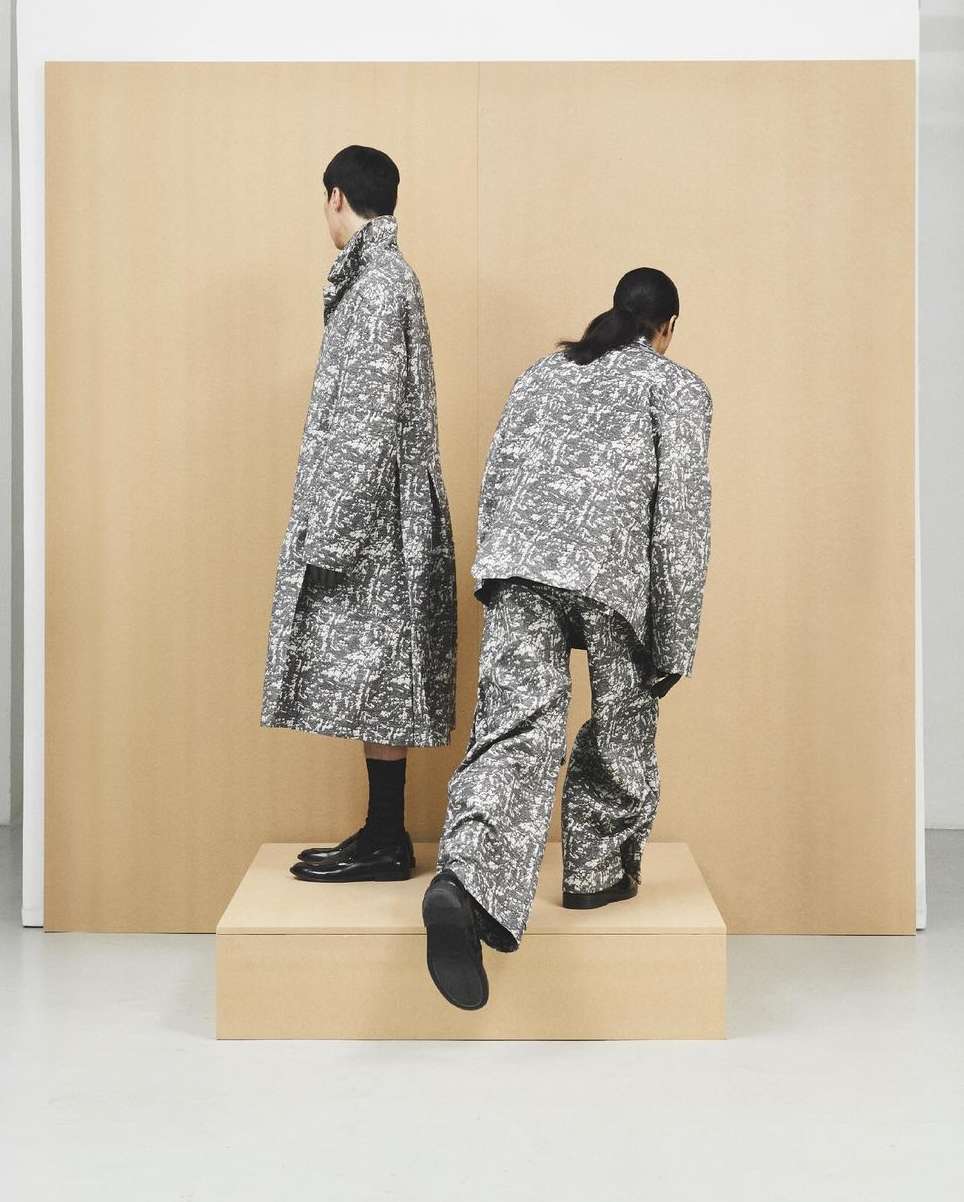 Two figures showcase grey and white patterned fashion, one in a long coat, the other in a jacket and pants, on a raised platform.