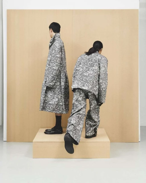 Two figures showcase grey and white patterned fashion, one in a long coat, the other in a jacket and pants, on a raised platform.