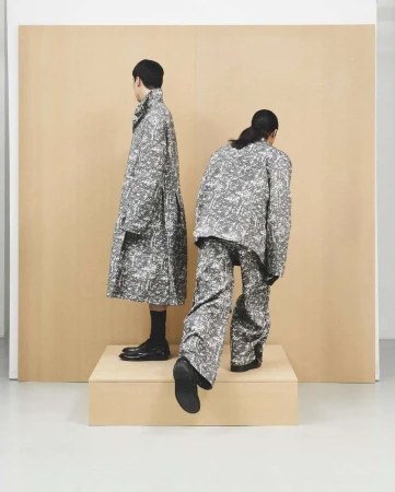 Two figures showcase grey and white patterned fashion, one in a long coat, the other in a jacket and pants, on a raised platform.