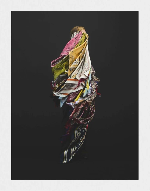 A woman stands against a dark background, draped in a cascade of colorful patterned scarves that obscure most of her figure.