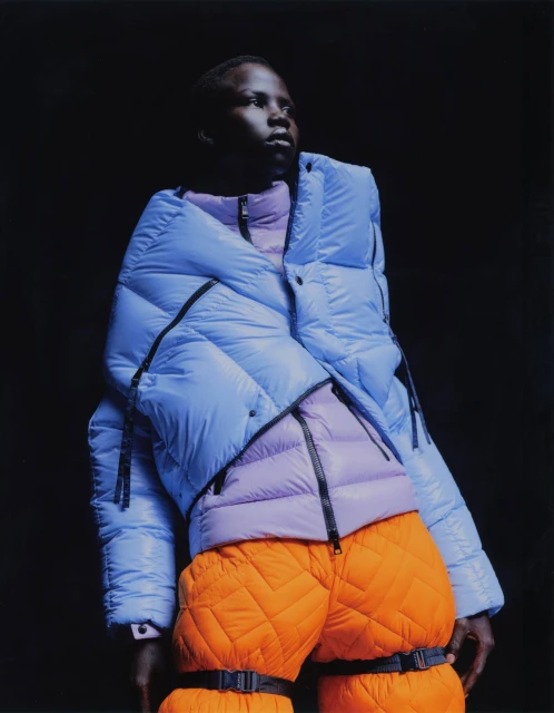 Fashion portrait of a Black adult male wearing a light blue puffer jacket over a lilac puffer vest, and orange quilted pants against a black backdrop.
