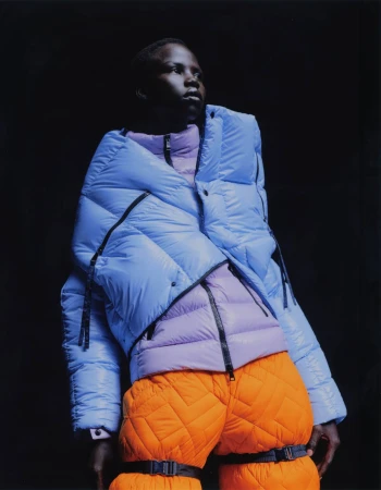 Fashion portrait of a Black adult male wearing a light blue puffer jacket over a lilac puffer vest, and orange quilted pants against a black backdrop.