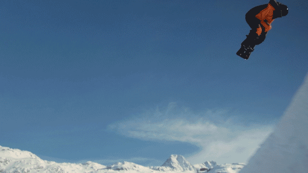 A snowboarder in mid-air against a blue sky, captured in a dynamic action shot. Snowy mountains are visible in the background.