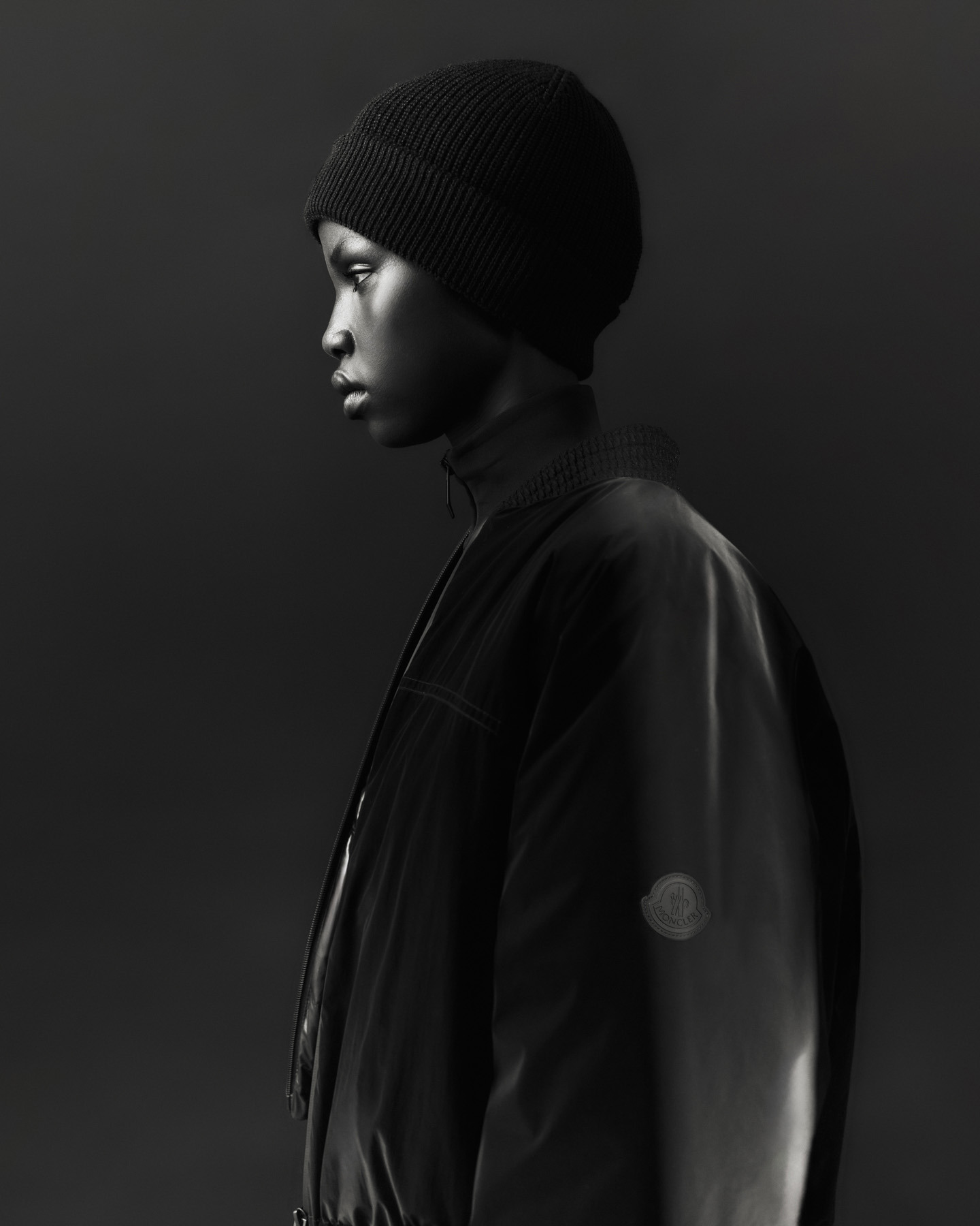 Monochrome portrait of a young adult in black beanie and jacket, captured in a photo studio setting with minimalist lighting.