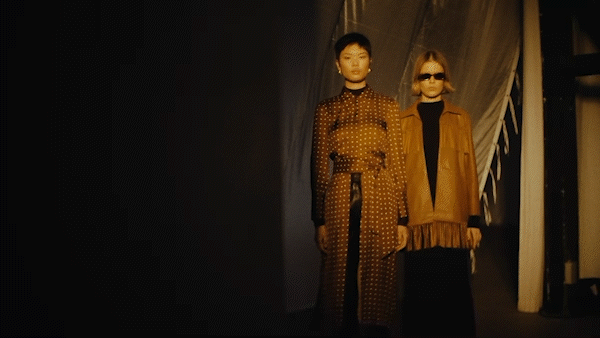 Two fashion models showcase apparel with an amber color palette in a dimly lit studio with a partial backdrop and modern apparel.