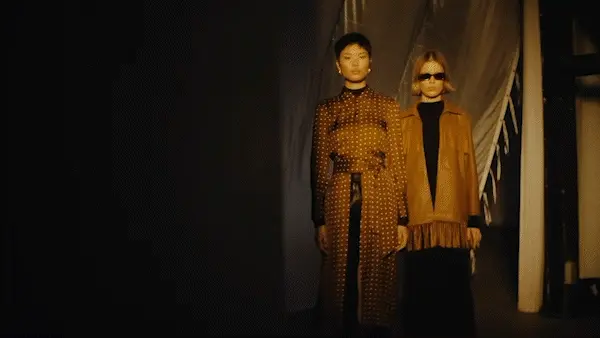 Two fashion models showcase apparel with an amber color palette in a dimly lit studio with a partial backdrop and modern apparel.
