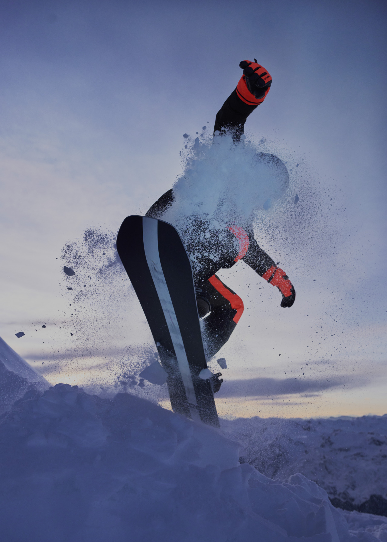 Snowboarder in mid-air performing trick on a black snowboard with snow spraying in the background during sunset in a wintry landscape.