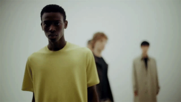 A dark-skinned male model in yellow T-shirt stands out against the white backdrop, with two blurred figures behind him in a fashion setting.