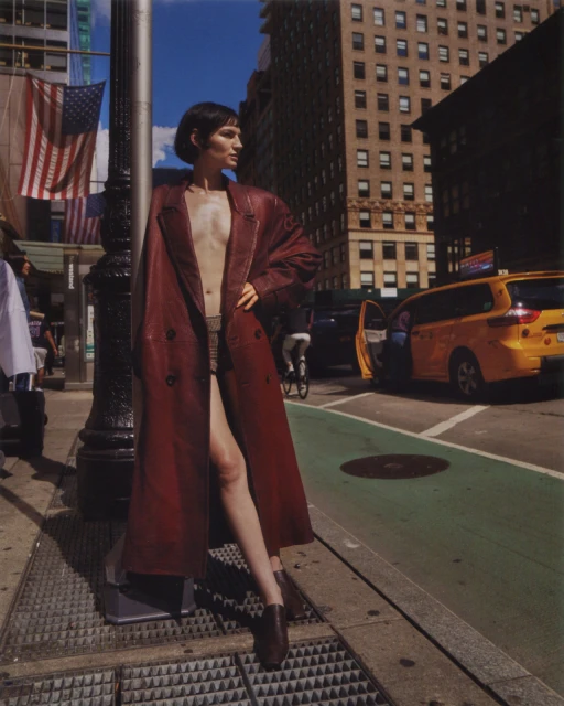 A fashionable woman in a long coat stands on a city street with tall buildings and a yellow taxi in the background on a sunny day.