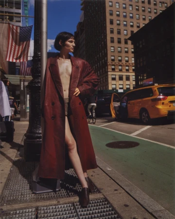 A fashionable woman in a long coat stands on a city street with tall buildings and a yellow taxi in the background on a sunny day.