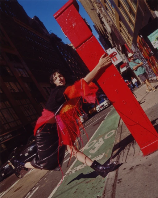 A young adult model stands on a city street in a black top, leather skirt, and bright, colorful fringed garment, striking a fashion pose.