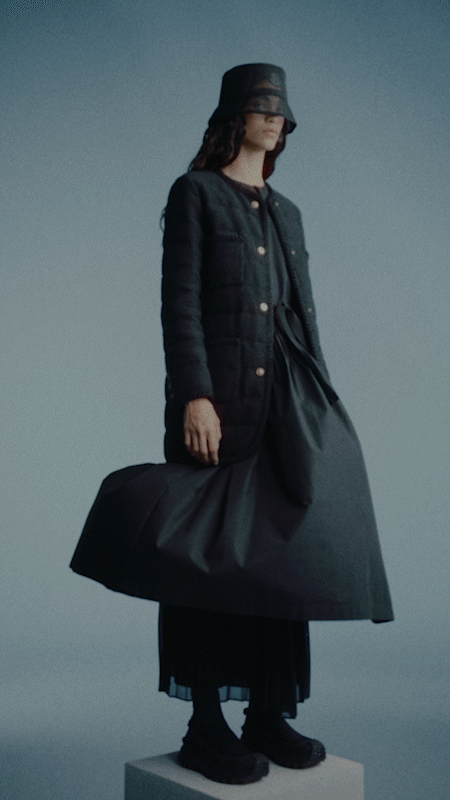 Fashionable adult woman poses in a jacket, skirt, and bucket hat against a plain background in a studio-lit setting.