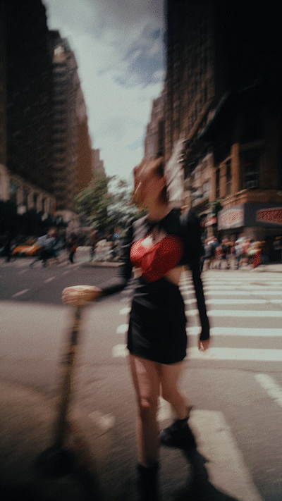 A young woman in a black dress and red top walks across a crosswalk in an urban street setting on a sunny day with buildings visible.
