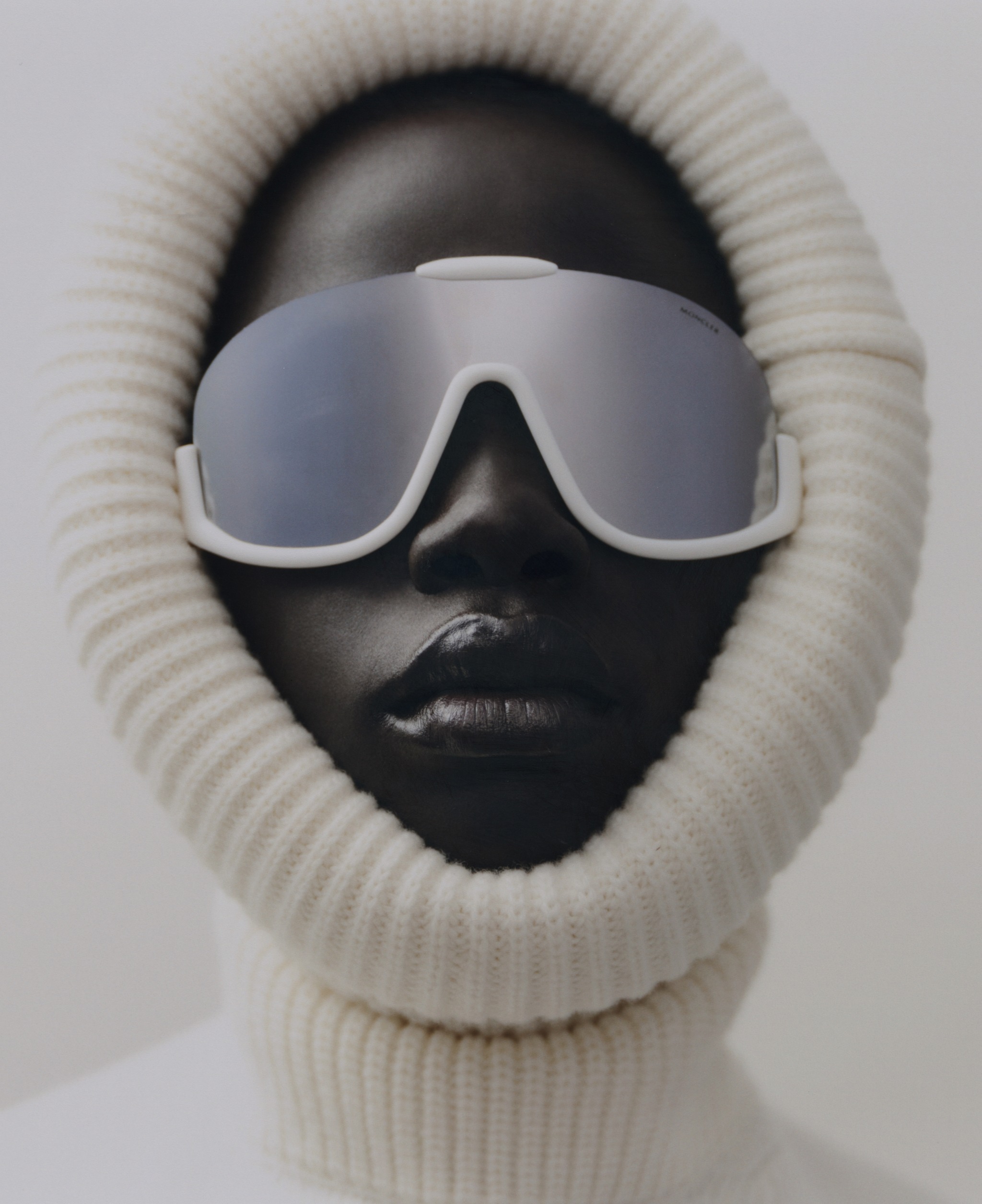 Close-up portrait of a Black woman with sunglasses and a knitted balaclava against a pale gray background. Fashionable and minimal aesthetic.