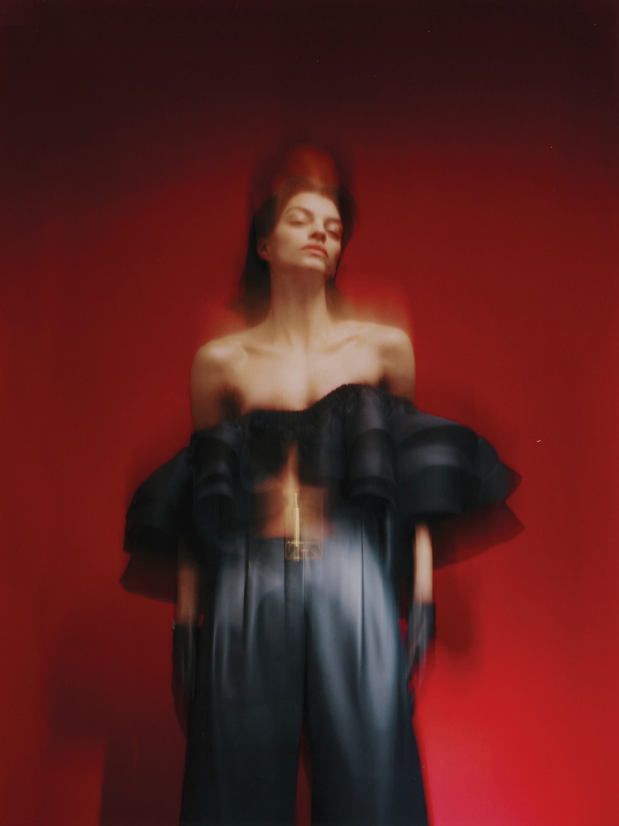 Fashion portrait of a female model wearing a black, off-the-shoulder top and wide-leg pants against a vibrant red background.