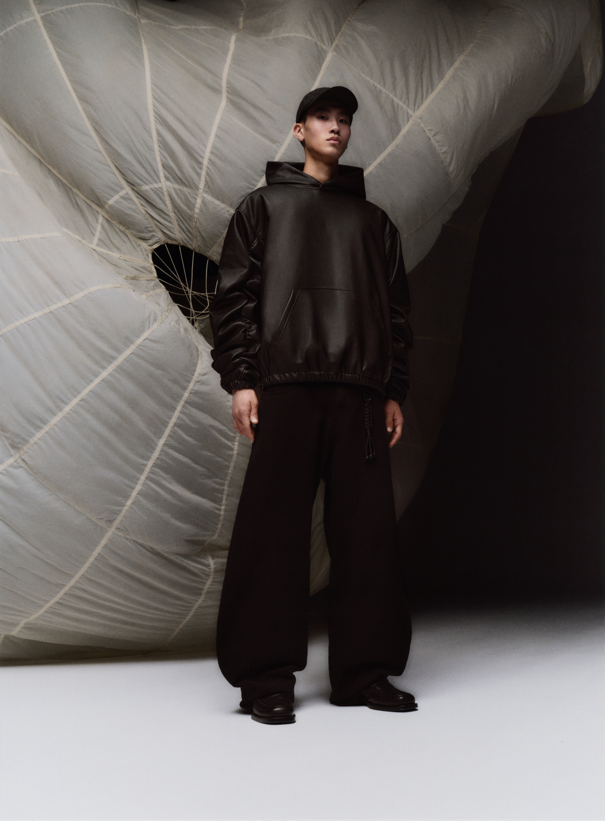 A person in a black hoodie, wide leg pants, and cap poses in a studio with a large white parachute-like object in the background.