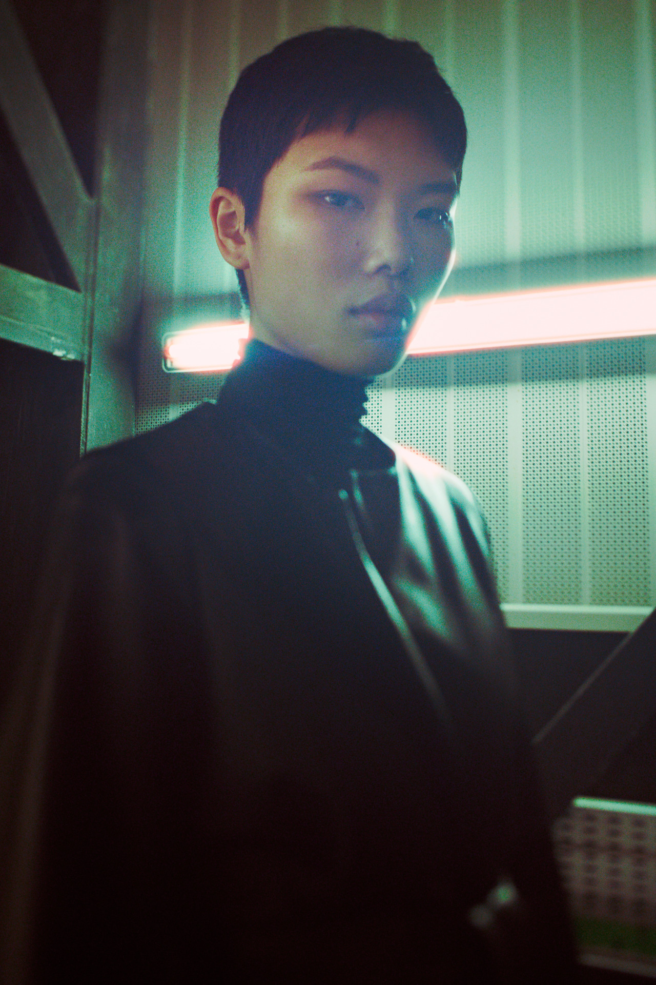 Portrait of a young adult with short hair wearing a dark turtleneck sweater and jacket indoors with light-green and gray walls.