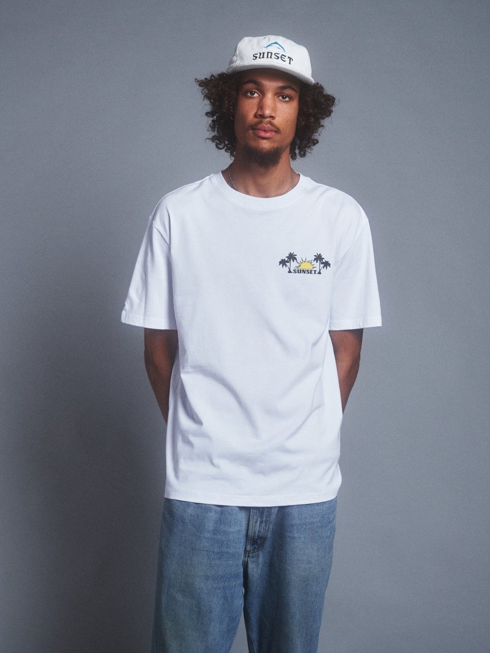 A curly-haired man wears a white t-shirt and hat with a palm tree graphic against a gray background in a studio setting. Medium shot.