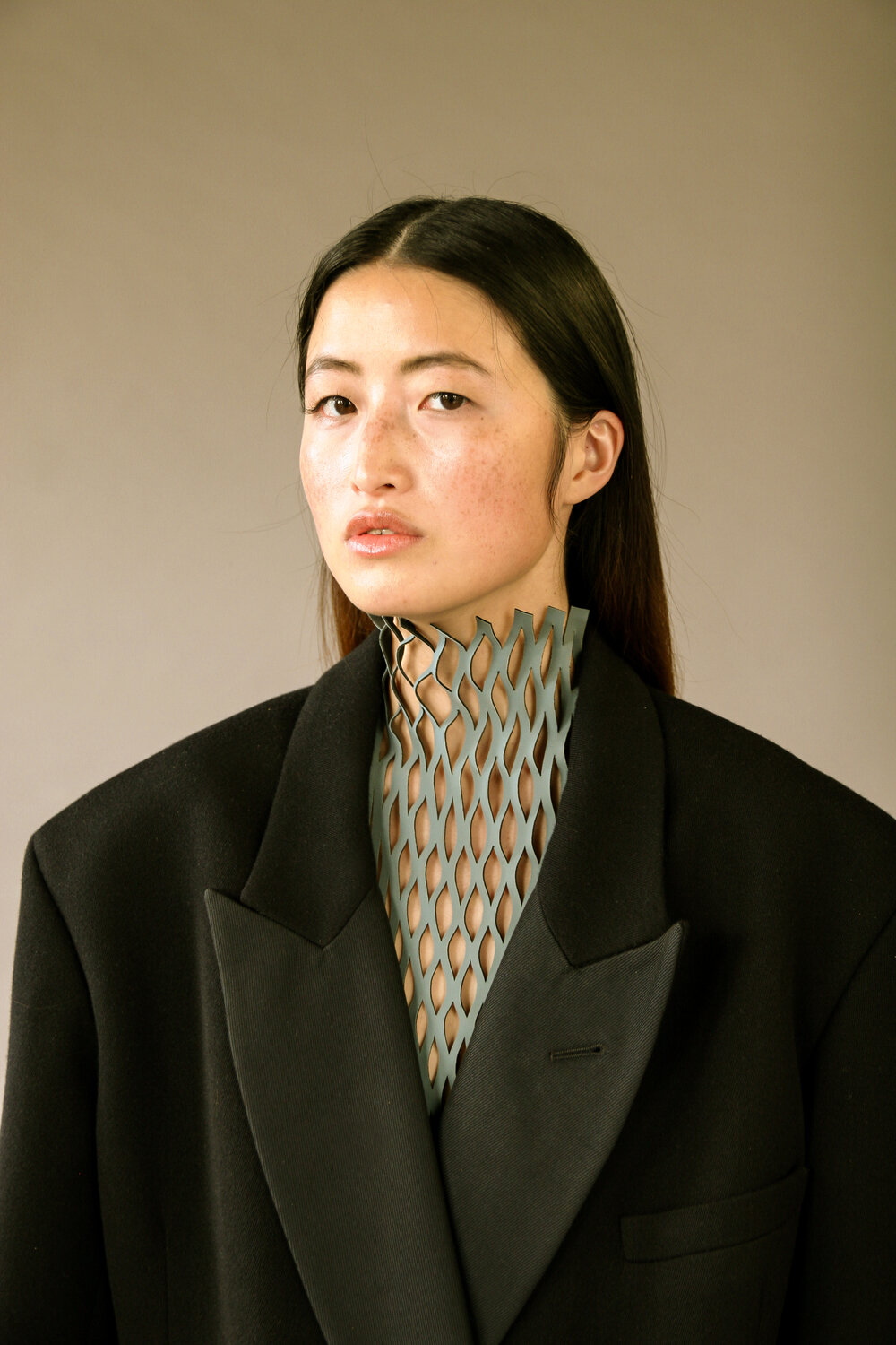 Portrait of an Asian woman wearing a black jacket and grey patterned high-neck garment against a neutral studio backdrop in soft, diffused light.