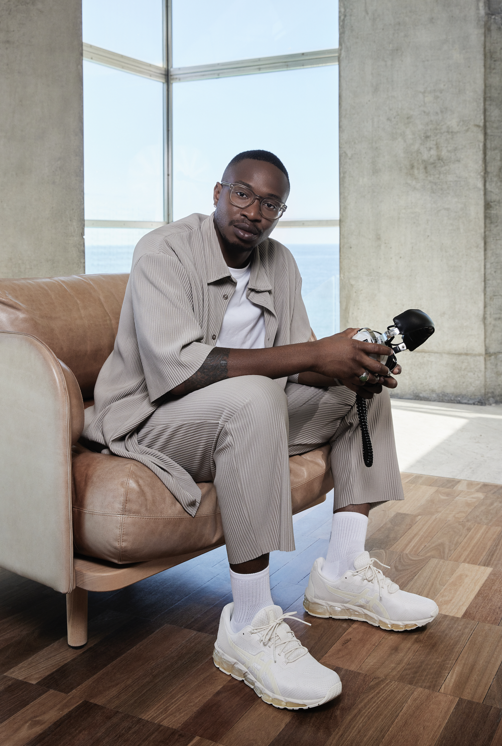 Indoor portrait of adult male in tan pleated outfit sitting in chair, wearing glasses and holding vintage camera, with light-colored sneakers.