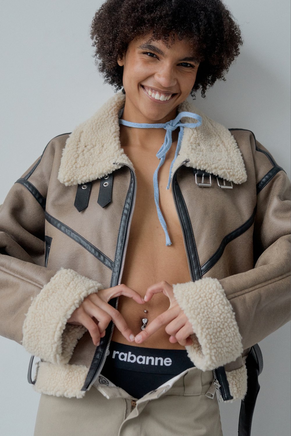 Smiling woman poses in a studio with a heart shape made by her hands, wearing a fashionable shearling jacket and designer underwear.
