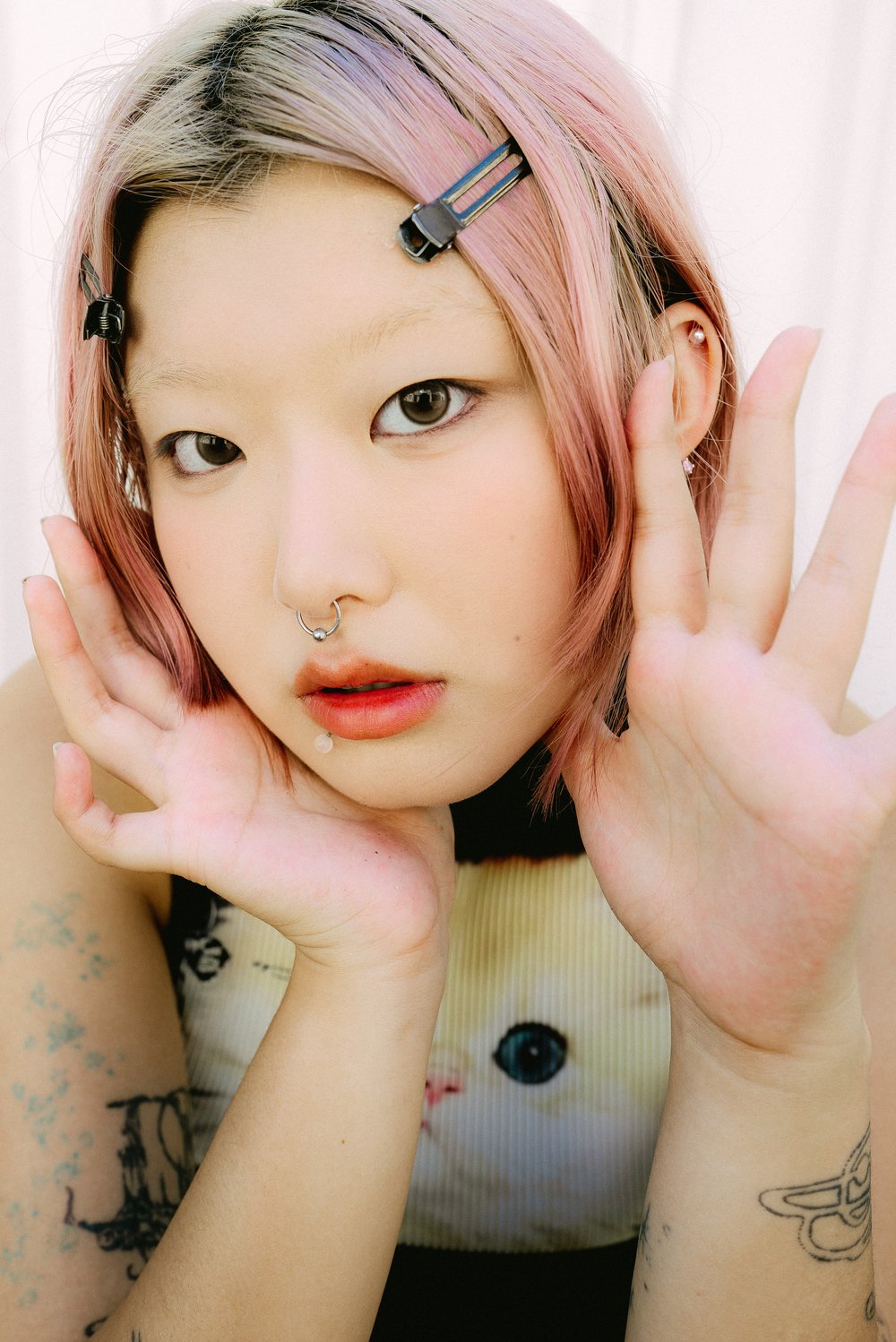 Close-up portrait of an Asian woman with pink hair and facial piercings, gesturing with her hands, wearing a top with a cat design.