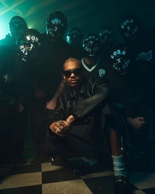Male musician wearing sunglasses poses with backup performers in masks on a black and white checkerboard floor with dark, moody lighting.