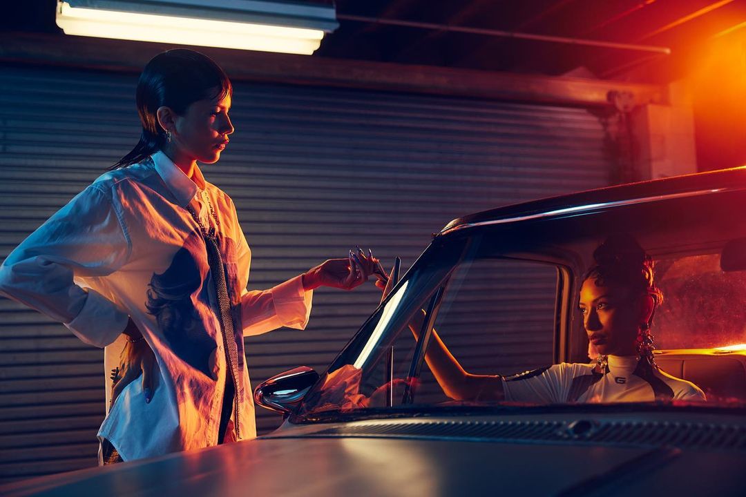 Two women in a garage with a vintage car. One stands next to the car while the other sits inside. Soft, atmospheric lighting.