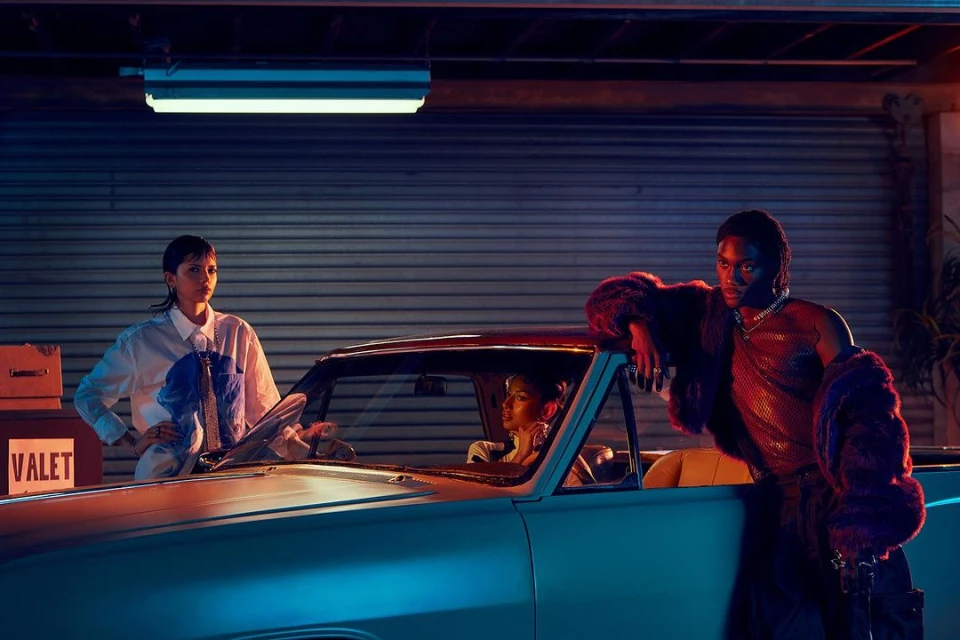 Three people pose beside a vintage car inside a garage with blue lighting, wearing stylish clothing and jewelry.