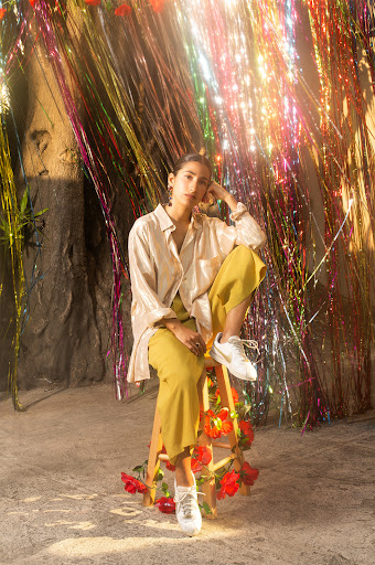 Woman seated on a stool wearing a white shirt and yellow trousers against a backdrop of metallic tinsel and red flowers