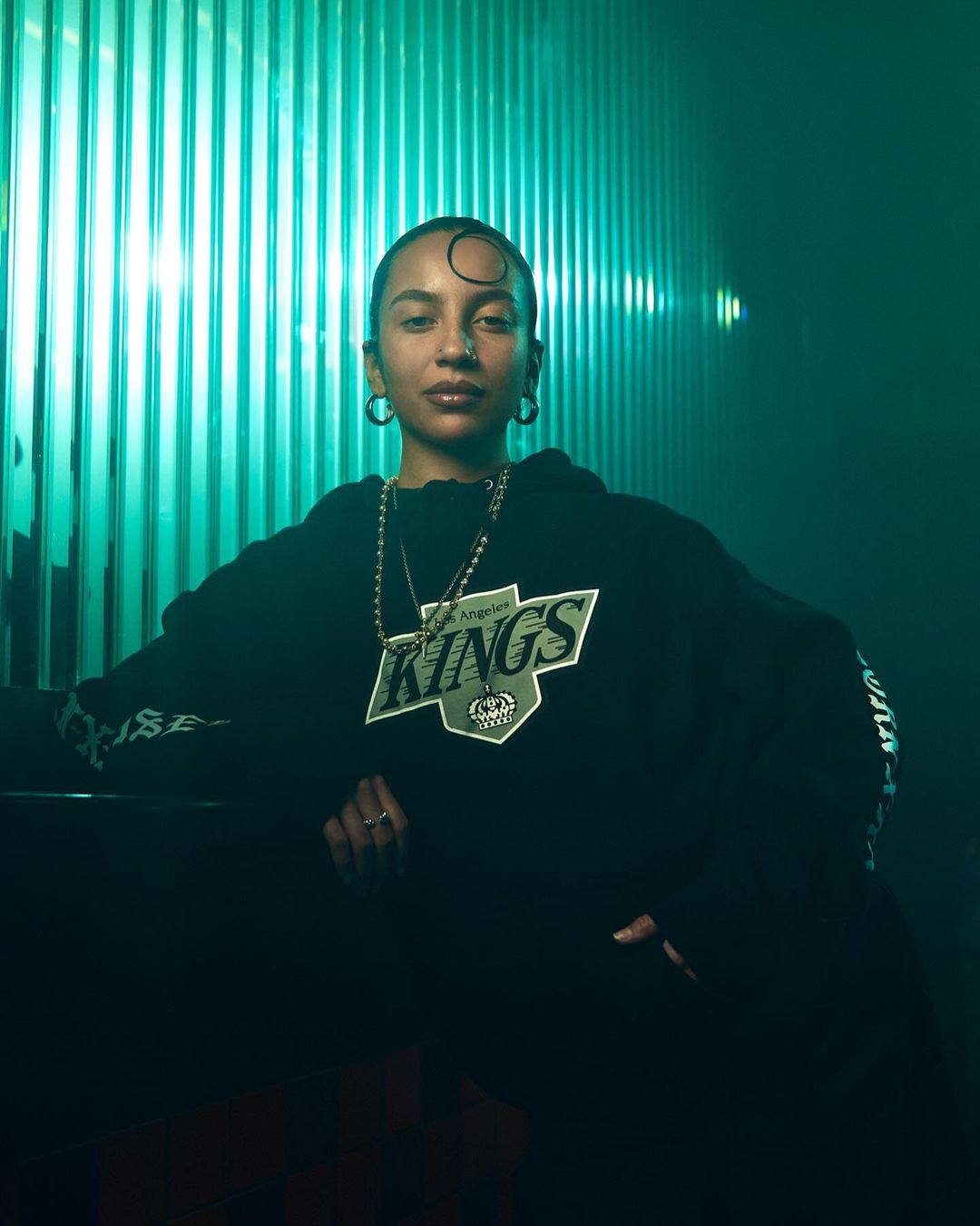 Portrait of a woman wearing a Los Angeles Kings hoodie in front of a turquoise corrugated metal wall, illuminated with studio lighting.