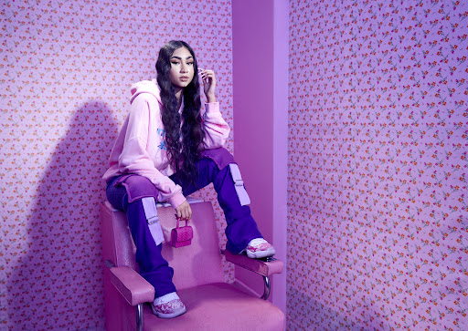 Young Asian woman in pink hoodie and purple pants sitting on a pink chair holding a miniature purse in a pink interior.