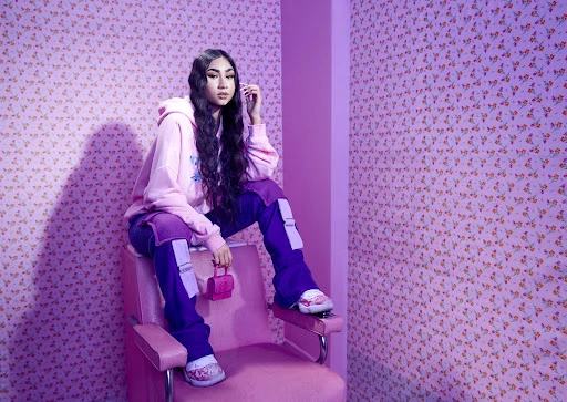 Young Asian woman in pink hoodie and purple pants sitting on a pink chair holding a miniature purse in a pink interior.