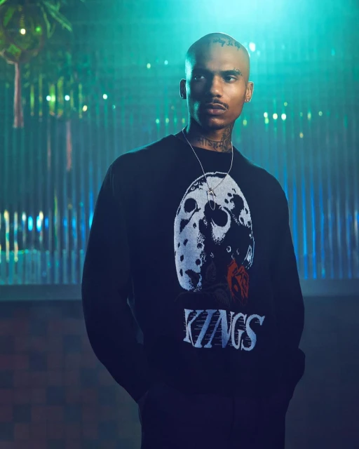 Portrait of an adult male in a black sweater with graphic. Indoor shot with neon lighting highlights facial tattoos and jewelry.